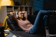 © cherryandbees - young woman relaxing on sofa at home. she is listening music on headphones . night evening scene