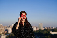 Portrait Of Cute Brunette Superhero Girl In Black Dress And Red Face Mask On Blue Sky And Urban City Background. Female Power, Women Rights, Protest Or Activism Concept. High Quality Image