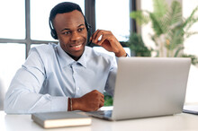 Successful Pleasant African American Guy In Headphones, Hotline Operator, Support Worker, Online Consultant Is Sitting At A Laptop In The Office, Talking To A Client On A Video Call, Smiling Friendly