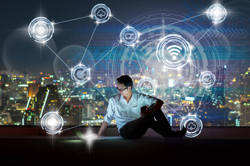 Wall Mural - Asian businessman sitting and using the laptop showing Wireless communication connecting of smart city Internet of Things Technology over the cityscape background, technology and innovation concept