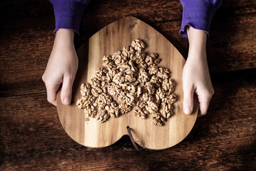 Wall Mural - Female hands hold a walnut. Peeled walnut halves on a wooden tray in the shape of a heart. Dark wooden background. Healthy eating. Vegetarianism.