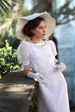 Young Woman In White Edwardian Style Dress And White Big Hat Standing In Old Abandoned Castle. Portrait Of Aristocratic Girl.