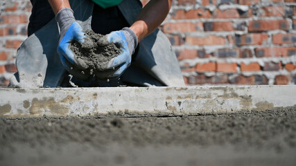 Close up of man hands in work gloves holding sand-cement mix for floor screed. Male worker preparing floor screed material while kneeling near screed rail at construction site.