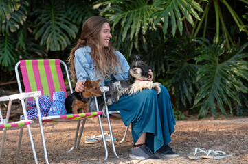 One woman sitting on a beach chair with a lhasa apso on her lap and one mixed breed dog on another chair by her side, on the beach, plants in the background, during the day. 