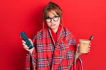 Poster - Redhead young woman wrapped in a red warm red blanket eating icecream and watching tv depressed and worry for distress, crying angry and afraid. sad expression.