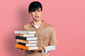 Poster - Handsome hipster young man holding a pile of books clueless and confused expression. doubt concept.