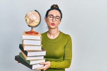 Wall Mural - Young hispanic girl holding a pile of books and world ball looking at the camera blowing a kiss being lovely and sexy. love expression.