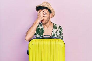 Wall Mural - Young hispanic man wearing summer style and holding cabin bag peeking in shock covering face and eyes with hand, looking through fingers afraid