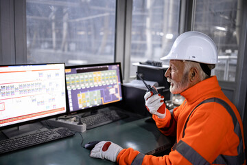 Wall Mural - Industrial worker in safety equipment sitting in factory control room monitoring production process and talking on radio communication.