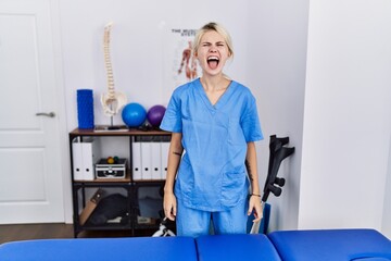 Wall Mural - Young physiotherapist woman working at pain recovery clinic angry and mad screaming frustrated and furious, shouting with anger. rage and aggressive concept.