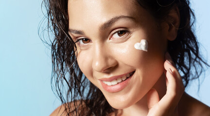 Pretty  girl with heart shaped face cream or foam on cheek, healthy skin, smiling during daily skincare routine, looking at camera on blue studio background. Natural woman beauty concept.