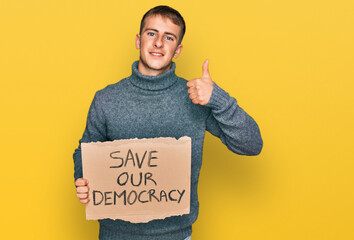 Wall Mural - Young blond man holding save our democracy protest banner smiling happy and positive, thumb up doing excellent and approval sign