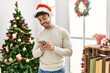 © Krakenimages.com - Young hispanic man wearing christmas hat using smartphone at home.