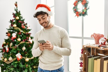 young hispanic man wearing christmas hat using smartphone at home.