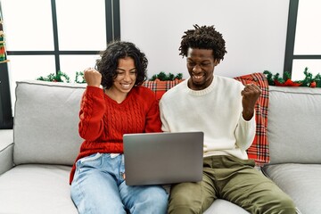 Poster - Young interracial couple sitting on the sofa on christmas using laptop screaming proud, celebrating victory and success very excited with raised arms