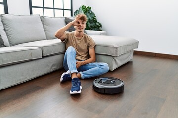 Sticker - Young caucasian man sitting at home by vacuum robot smiling happy doing ok sign with hand on eye looking through fingers