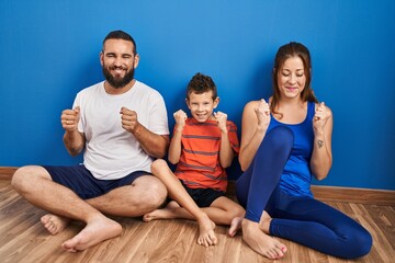 Wall Mural - Family of three sitting on the floor at home excited for success with arms raised and eyes closed celebrating victory smiling. winner concept.