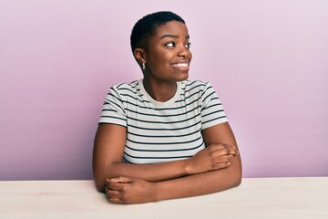 Sticker - Young african american woman wearing casual clothes sitting on the table looking away to side with smile on face, natural expression. laughing confident.