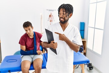 Canvas Print - Young hispanic man working at pain recovery clinic with a man with broken arm pointing aside worried and nervous with forefinger, concerned and surprised expression