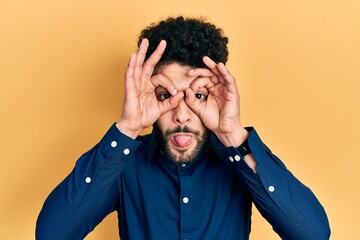 Sticker - Young arab man with beard wearing casual shirt doing ok gesture like binoculars sticking tongue out, eyes looking through fingers. crazy expression.