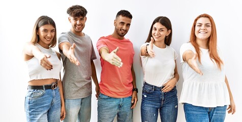 Poster - Group of young friends standing together over isolated background smiling friendly offering handshake as greeting and welcoming. successful business.