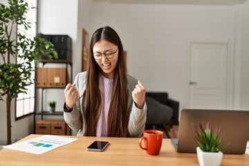 Sticker - Young chinese business worker wearing business style sitting on desk at office very happy and excited doing winner gesture with arms raised, smiling and screaming for success. celebration concept.