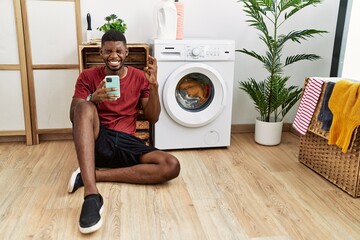 Poster - Young african american man using smartphone waiting for washing machine gesturing finger crossed smiling with hope and eyes closed. luck and superstitious concept.