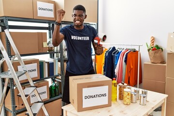 Poster - Young african american volunteer man packing donations box for charity angry and mad raising fist frustrated and furious while shouting with anger. rage and aggressive concept.