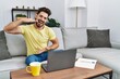 © Krakenimages.com - Young man with beard using laptop at home gesturing with hands showing big and large size sign, measure symbol. smiling looking at the camera. measuring concept.