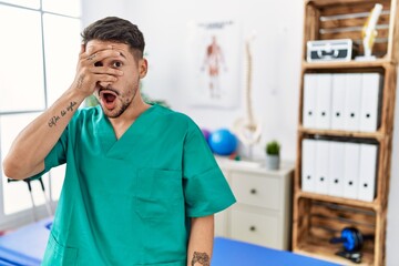 Canvas Print - Young physiotherapist man working at pain recovery clinic peeking in shock covering face and eyes with hand, looking through fingers with embarrassed expression.