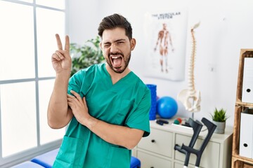 Poster - Young man with beard working at pain recovery clinic smiling with happy face winking at the camera doing victory sign. number two.