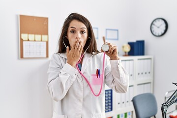 Poster - Young doctor woman wearing doctor coat holding stethoscope covering mouth with hand, shocked and afraid for mistake. surprised expression