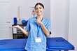© Krakenimages.com - Young hispanic woman wearing physiotherapist uniform standing at clinic amazed and smiling to the camera while presenting with hand and pointing with finger.