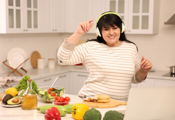 Poster - Happy overweight woman with headphones dancing while cooking in kitchen