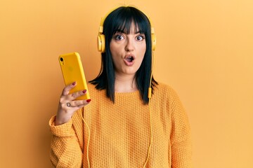 Poster - Young hispanic woman using smartphone wearing headphones scared and amazed with open mouth for surprise, disbelief face