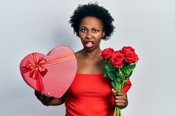 Sticker - Young african american woman holding anniversary present and bouquet of flowers in shock face, looking skeptical and sarcastic, surprised with open mouth