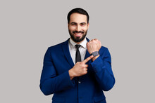 Good Result And On Time. Positive Bearded Man Standing, Showing His Smart Watch And Looking At Camera With Toothy Smile, Wearing Official Style Suit. Indoor Studio Shot Isolated On Gray Background.