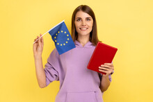 Portrait Of Smiling Beautiful Student Woman Holding Books And Europe Union Flag, Education Abroad, Wearing Purple Hoodie. Indoor Studio Shot Isolated On Yellow Background.