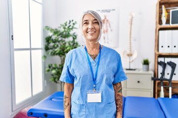 Poster - Middle age grey-haired woman wearing physiotherapist uniform at medical clinic with a happy and cool smile on face. lucky person.