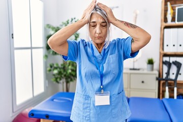 Poster - Middle age grey-haired woman wearing physiotherapist uniform at medical clinic suffering from headache desperate and stressed because pain and migraine. hands on head.