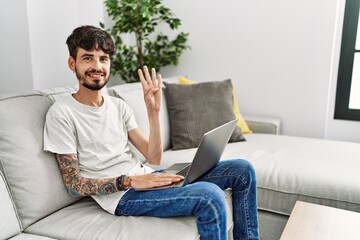 Poster - Hispanic man with beard sitting on the sofa showing and pointing up with fingers number four while smiling confident and happy.