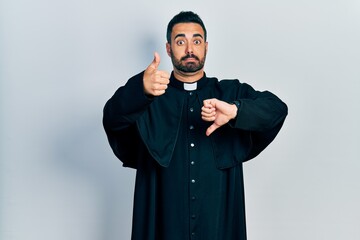 Canvas Print - Handsome hispanic man with beard wearing catholic priest robe doing thumbs up and down, disagreement and agreement expression. crazy conflict