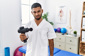 Wall Mural - Young indian physiotherapist holding therapy massage gun at wellness center in shock face, looking skeptical and sarcastic, surprised with open mouth