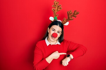 Poster - Young hispanic woman wearing deer christmas hat and red nose in hurry pointing to watch time, impatience, upset and angry for deadline delay