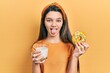 Young brunette girl eating donut and drinking glass of milk sticking tongue out happy with funny expression.