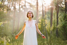 Unity with nature. Young woman in white nightgown standing in green forest on sunny morning with closed eyes and outstretched arms, female breathing deep fresh air in meadow at sunrise. Forest Bathing