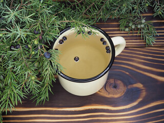 Herbal tea with juniper berries in an enamel mug, green twigs with fruits on a bright wooden background, top view, flat layout. Useful juniperus plant for use in medicine, cooking 