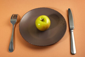 Wall Mural - The apple is on a brown plate on a light brown background.  The fork and knife are next to the plate. The concept of proper nutrition