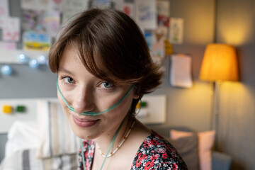 Face of smiling teenage girl with oxygen pipe in her nose looking at camera against interior of hospital ward