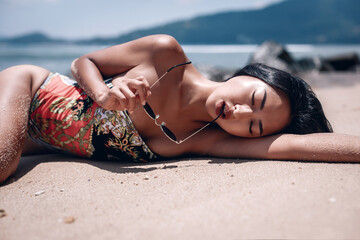 Girl with black hair and perfect body is lying on the sand holding sunglasses in her teeth